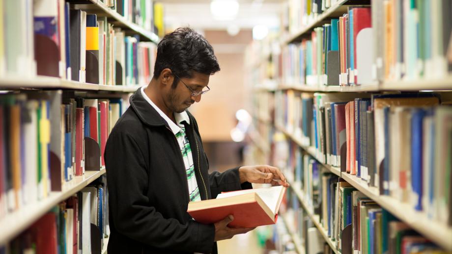 photo of a young man standing between two library shelves reading a book