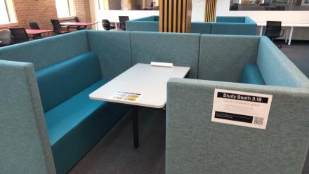 Bookable study booth in a library learning space, featuring teal upholstered bench seating on three sides around a central table with a power outlet. Similar study booths, desks, chairs, large windows, and a wall-mounted display screen are visible in the background. A sign on the booth reads “Study Booth 3.18”.